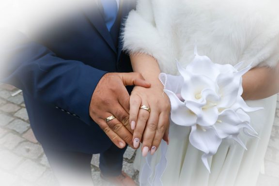 Hände eines Paares mit Hochzeitsringen, umgeben von einer weißen Blume.