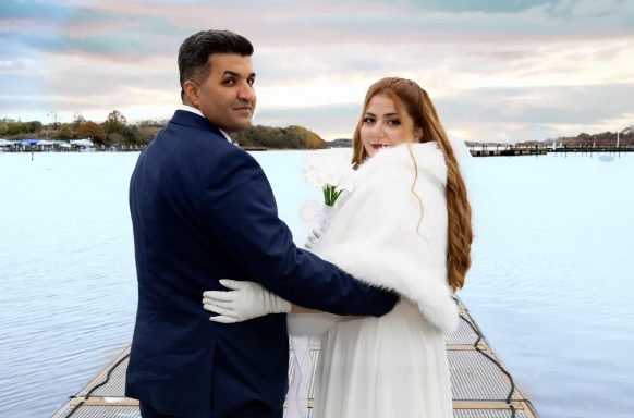 Ein Brautpaar steht am Wasser, der Mann im Anzug, die Frau in einem weißen Kleid mit Umhang.